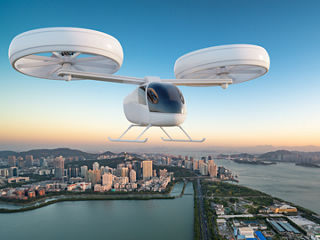 Electric vehicle takeoff and landing aircraft flying over a coastal city at sunrise, with high-rise buildings, water, and mountains in the background.