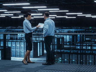 Two IT professionals, a man and a woman, stand on a walkway and discuss information on a tablet in a large, modern data center filled with server racks.