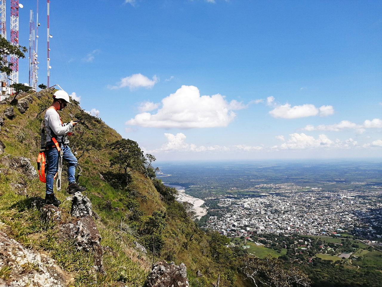 A field technician uses handheld test equipment near telecommunications towers on a ridge overlooking a city.