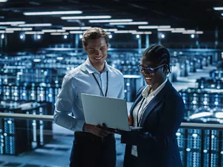Two engineers review information on a laptop while standing among rows of server racks in a data center.