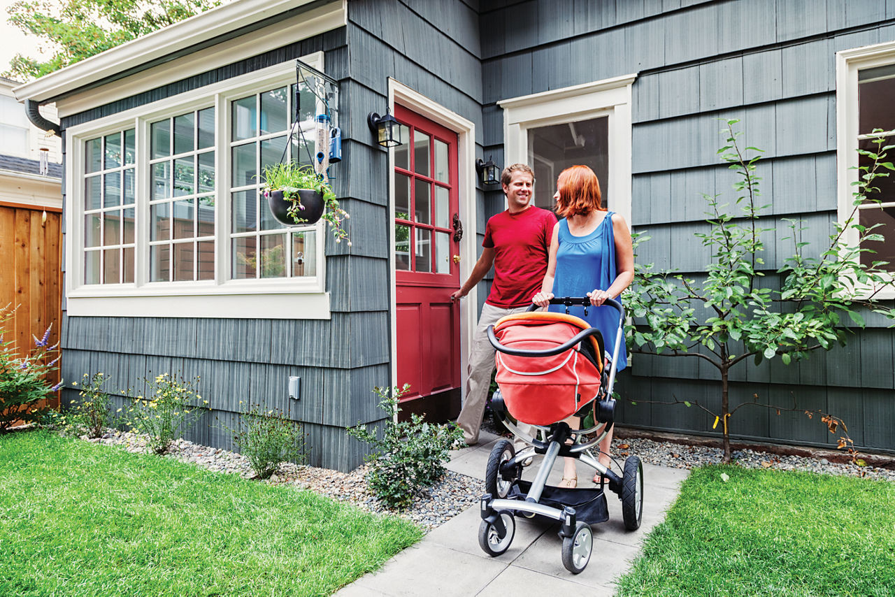 Photo of a young family on their way out to take a walk with their new baby in a stroller; husband is shutting the door behind him.