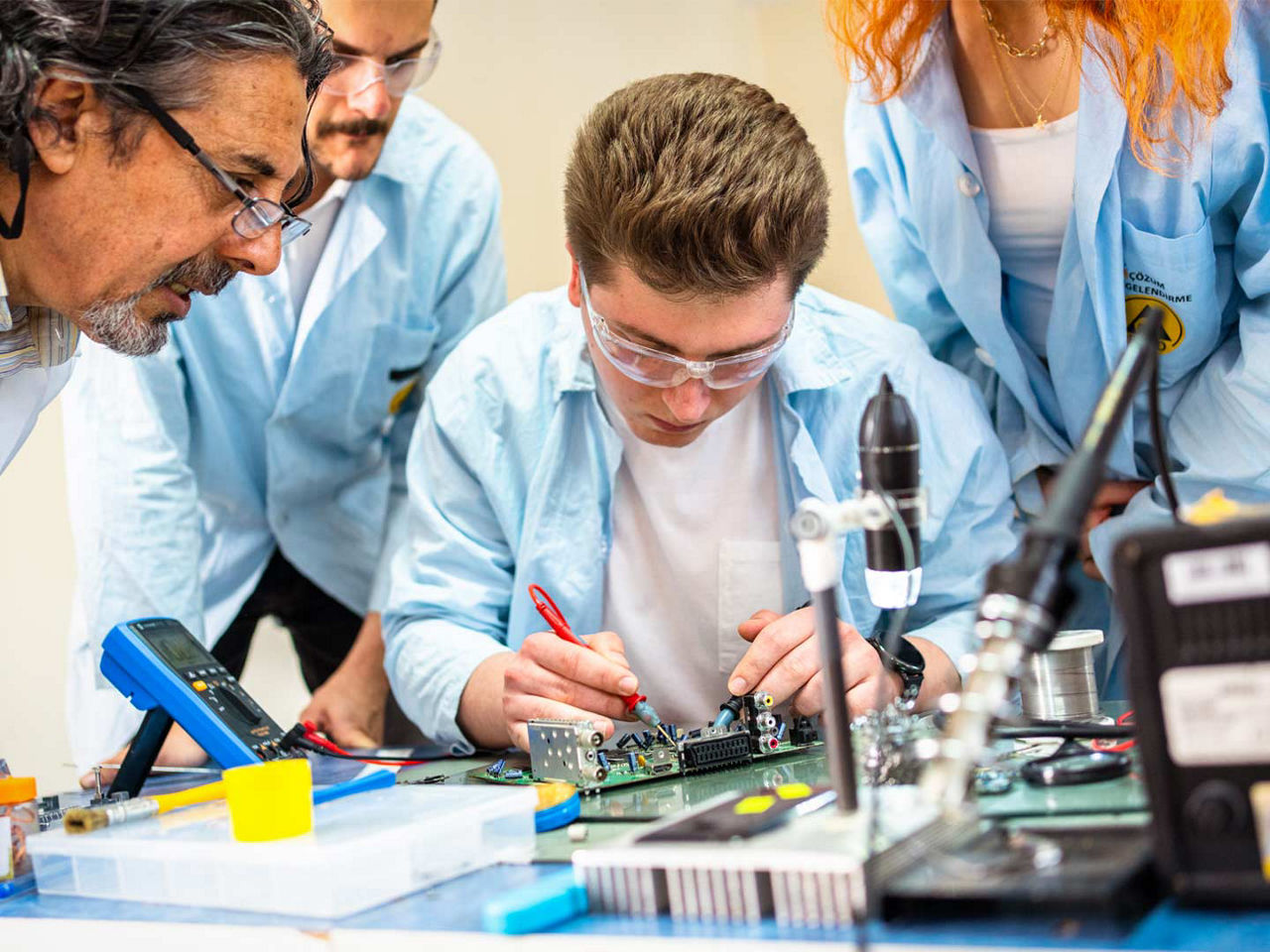 A technician working on a printed circuit board while several other individuals observe.