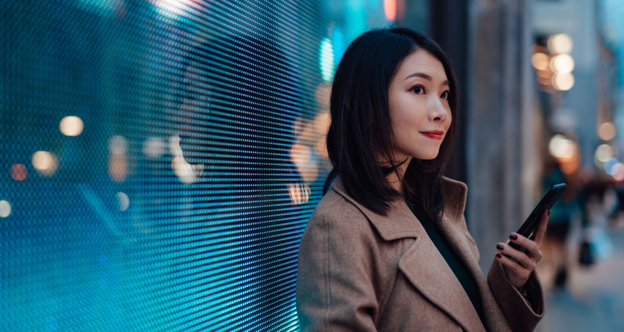 Young Asian business woman using mobile phone while waiting on the city street at night. Against a massive digital display with city lights illuminated at the background. Connect the future.
