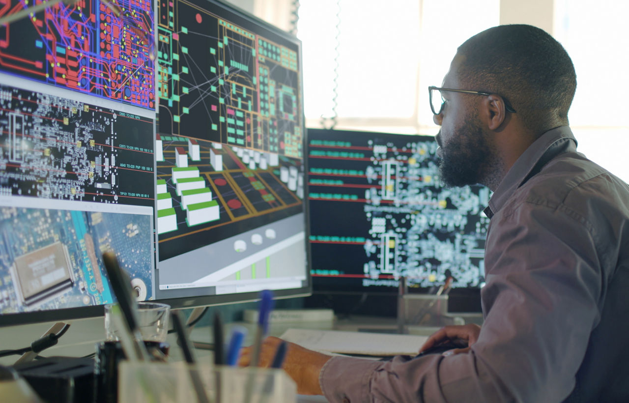 Stock image of an Afro-Caribbean male designing electronic circuit boards ( PCBs).&#xA;Heâ  s sitting at a desk with a large computer screen displaying a variety of apps & PCB illustrations.